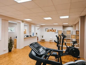 Salle d'accueil du cabinet de kinésithérapie Le Pôle avec le comptoir d'accueil, des chaises, et aussi un espace cardio composé d'un tapis de course, d'un vélo elliptique, d'un rameur et d'un vélo d'appartement.