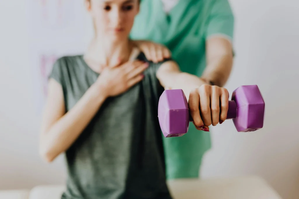 Une femme effectue des exercices de rééducation avec une haltères violette pour son épaule. Elle est assistée par son kinésithérapeute.