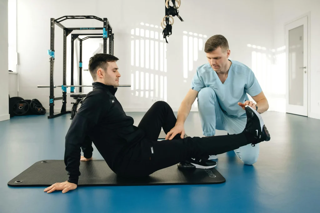 Un kiné accompagne un patient sportif pour effectuer un étirement de sa jambe droite. Le patient est assis sur un tapis d'exercice