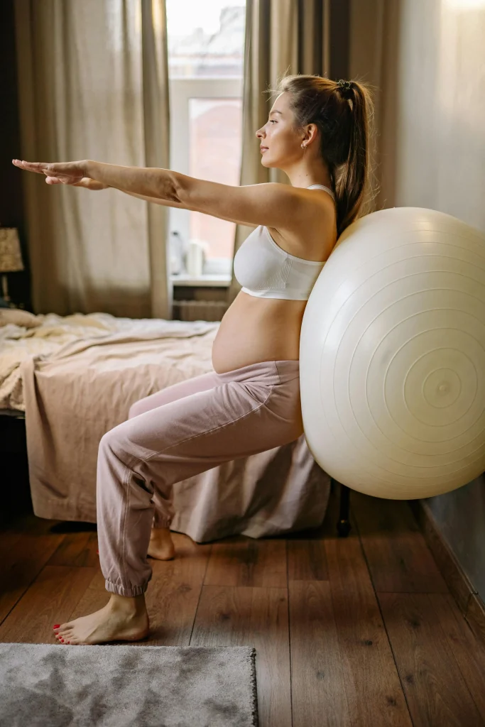 Une femme enceinte se tient adossée contre un mur en position chaise avec un ballon de fitness dans le dos.